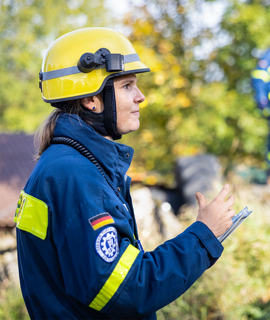 Anna Mirl im Profil, in blauer THW-Uniform und gelbem Schutzhelm, ein Klemmbrett in der Hand.